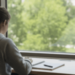 University student writing in a notebook at a desk beside a sunlit window